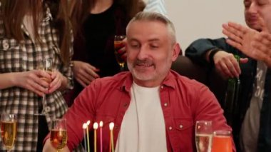 Şenlik şapkalı gri saçlı bir adam arkadaş çemberinde oturur ve pastadaki mumları söndürür. Ofis çalışanı doğum gününü ofiste iş arkadaşlarıyla kutlar. Yüksek kalite 4k görüntü