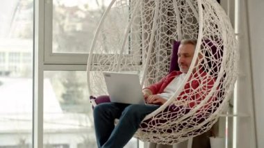 Kırmızı gömlekli bir adam koza sandalyesinde dizüstü bilgisayarıyla oturuyor ve daktilo ediyor. Gri saçlı bir adam pencerenin yanındaki sallanan sandalyede otururken dizüstü bilgisayarda çalışıyor. Yüksek kalite 4k görüntü