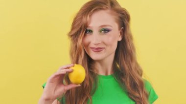 Kameraya bakıp elinde limon tutarken gülümseyen kızıl saçlı hoş bir kadın. Sarı duvara karşı kameraya poz veren gülümseyen kadın. Yüksek kalite 4k görüntü