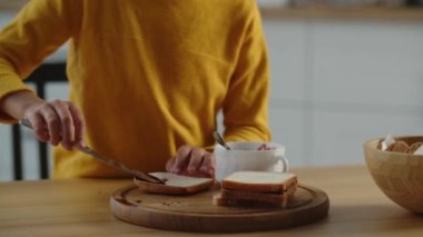 Mutfak masasında kendine sandviç yapan bir çocuğun görüntüsü. Sarı kazaklı bir çocuk mutfakta bir dilim ekmeğe ahududu reçeli sürüyor. Yüksek kalite 4k görüntü