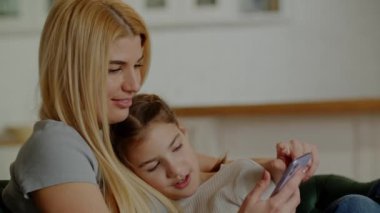 Mutlu bir aile, genç bir anne ve büyüyen bir kız telefona bakar. Sevimli küçük kız ve güzel genç annesi kanepede birlikte oturuyorlar, akıllı telefon kullanıyorlar ve gülümsüyorlar. Yüksek kalite 4k