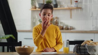 Sarı kazaklı bir çocuk mutfak masasının yanında duruyor ve bir elma yiyor. Küçük bir çocuk sulu olgun bir elmanın tadını çıkarıyor. Yüksek kalite 4k görüntü