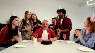 Beyaz saçlı, şenlikli şapkalı bir adam arkadaşlarıyla birlikte oturur ve pastadaki mumları söndürür. Ofis çalışanları iş arkadaşlarının doğum gününü kutluyorlar. Yüksek kalite 4k görüntü