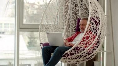 Kırmızı gömlekli bir adam koza sandalyesinde dizüstü bilgisayarıyla oturuyor ve daktilo ediyor. Gri saçlı bir adam pencerenin yanındaki sallanan sandalyede otururken dizüstü bilgisayarda çalışıyor. Yüksek kalite 4k görüntü