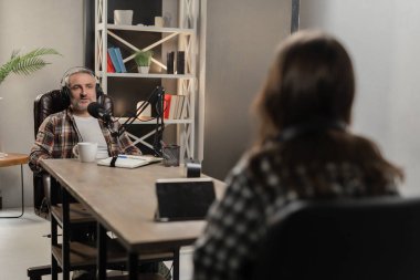 Kulaklıklı bir adam bir kadının karşısındaki masada oturur ve mikrofona bir şeyler söyler. İki blogcu stüdyodan ortak bir podcast kaydediyor. Yüksek kalite fotoğraf