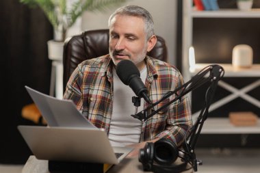 Sakallı bir adam bir dizüstü bilgisayarın ve mikrofonun önünde oturur ve bir kağıt parçasına bakar. Kayda başlamadan önce bir erkek blogcu yeni videosu için senaryoyu kontrol ediyor. Yüksek kalite fotoğraf