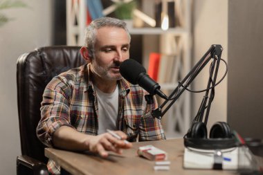 Elinde sigara olan bir adam bir masaya oturur ve stüdyo mikrofonuna bir şeyler söyler. Gri saçlı, sakallı bir adam masaya oturur ve bir podcast kaydeder. Yüksek kalite fotoğraf