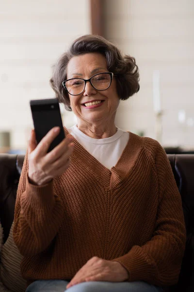 Sevimli yaşlı bayan, akıllı telefonundaki başvuruları kullanarak kanepede dinleniyor. İnternetten alışveriş yaparken arkadaşlarıyla ya da çocuklarıyla kurye ya da sosyal ağda sohbet eden mutlu, olgun bir kadın.