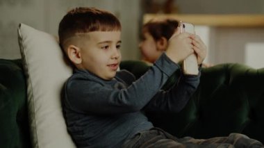  Kanepede oturan küçük çocuk telefonuyla eğleniyor. Arka plandaki bebek gülüp eğleniyor. Yüksek kalite 4k görüntü