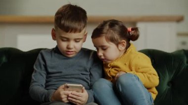 Kardeşler rahat koltukta otururken modern telefonda oyun oynuyorlar. Çocuklar akıllı telefondan sanal oyun oynuyorlar, internette komik içerikler izliyorlar. Yüksek kalite 4k görüntü