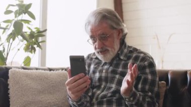 Video görüşmesi sırasında telefon ekranına bakan kıdemli adam. Gözlüklü ve kareli gömlekli büyükbaba telefonda konuşuyor. Yüksek kalite 4k görüntü