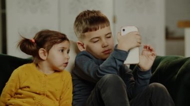 Odaklanmış küçük çocuklar koltukta oturup ailelerinin kontrolü olmadan akıllı telefonlarla oynuyorlar. Çocuklar ve elektronik cihazlar. Yüksek kalite 4k görüntü