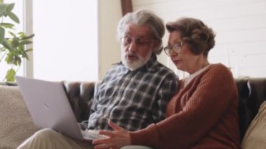 Kanepeye dizüstü bilgisayar kullanan orta yaşlı bir çift. Beyaz saçlı. Yaşlılar için yeni teknoloji. Yüksek kalite 4k görüntü