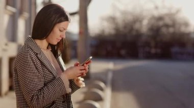 Çekici bir esmer yolun kenarında duruyor ve telefonuna bir şeyler yazıyor. Otobüs durağındaki bir kız. Yüksek kalite 4k görüntü