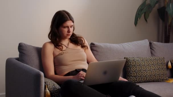 一个年轻的女人坐在沙发上 膝盖上放着笔记本电脑 在网上冲浪 一个漂亮的黑发女人坐在家里的沙发上 一边在笔记本电脑上打字 优质Fullhd影片 — 图库视频影像