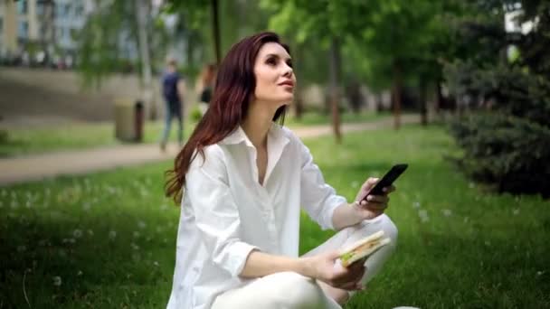 饥饿的女孩在城市公园吃三明治 享受美味的食物 女人在午休时间吃饭 并坐在社交网络上 速食概念 是的高质量的4K镜头 — 图库视频影像