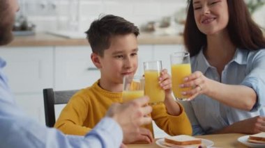 Oğlu portakal suyuyla bardakları tokuşturan gülümseyen aile. Çocuk ebeveynleriyle kahvaltıyı seviyor. Yüksek kalite 4k görüntü