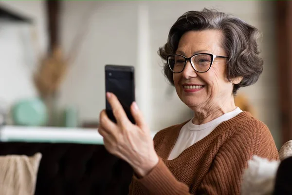 Happy mature middle aged woman enjoying using mobile apps, texting, sitting on sofa, smiling senior woman holding smart phone, looking at mobile phone screen, browsing social media at home. High