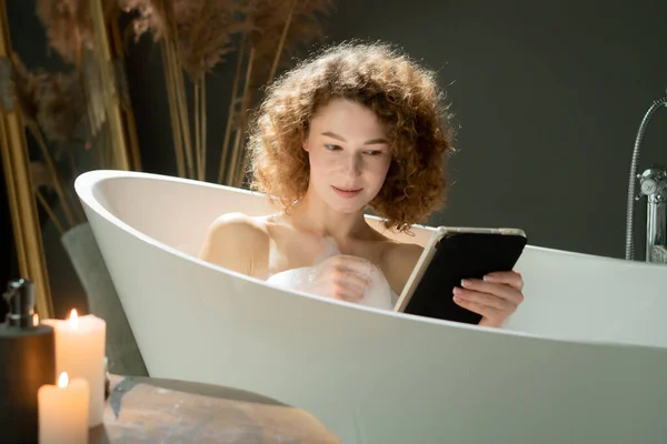 魅力的な若い女性がお風呂に横たわり デジタルタブレットで何かを見ています 美しい女性は夜の風呂に入っている間 彼女のタブレットにソーシャルメディアを閲覧しています 高品質の写真 — ストック写真