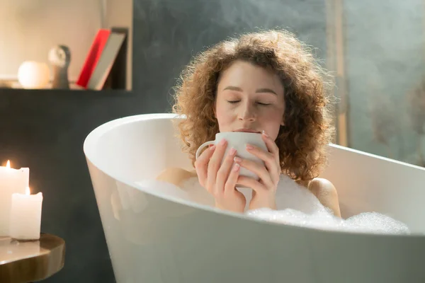 快乐的年轻女人睡觉前洗个澡喝茶 一个卷发的女人在泡泡浴中放松下来 喝甘菊茶来缓解压力 高质量的照片 — 图库照片