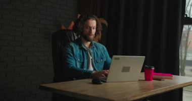 Şık, rahat bir oturma odasında siyah deri bir sandalyede otururken dizüstü bilgisayarda çalışan konsantre beyaz bir adam. Genç bir serbest çalışan evden çalışır. İnternette geziniyor, kahve içiyor.