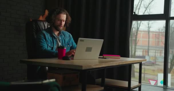 专注于笔记本电脑上工作的年轻人坐在时尚办公室的黑色皮椅上 一个英俊的自由职业者在家里工作 浏览互联网 喝纸杯咖啡 — 图库视频影像