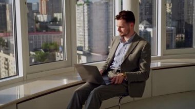Professional Creative Man Sitting on chair in Office Studio Working on a Laptop. He is focused on work and inspired by the beauty of the city on background. High quality 4k footage