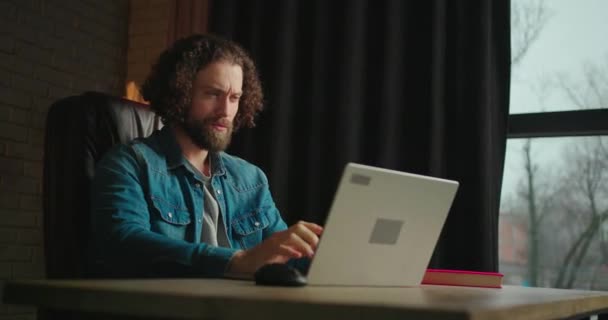 スタイリッシュなオフィスで黒い革の椅子に座っている間 ラップトップコンピュータで働くことに集中した若い男 男は仕事を解決することを考え 顔に触れる 若いフリーランサーの作品 — ストック動画