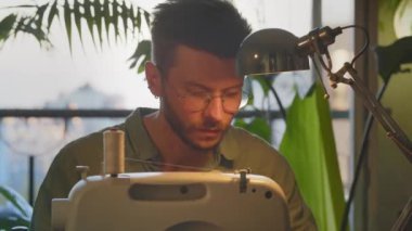 Erkek giysi tasarımcısı evden dikiş makinesinde çalışıyor. Genç girişimci, evde büyük yeşil bitkilerle iş yapmaya başlıyor. El işi ve imalat kavramı. Yüksek kalite 4k