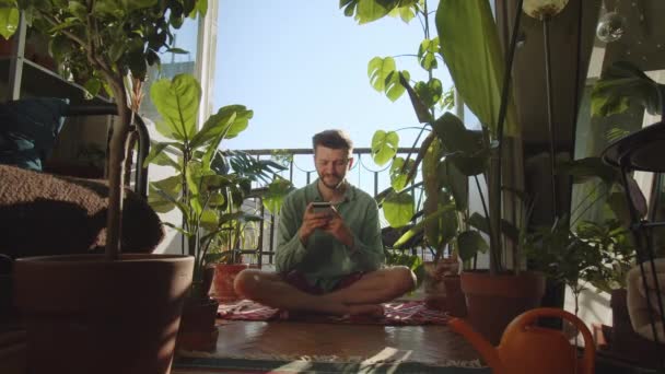 一个穿着绿色T恤 手里拿着绿色手机的年轻嬉皮士用智能手机留言 房间里的绿色植物是生态型的 和平与幸福 生态活动家 生态行动主义 — 图库视频影像