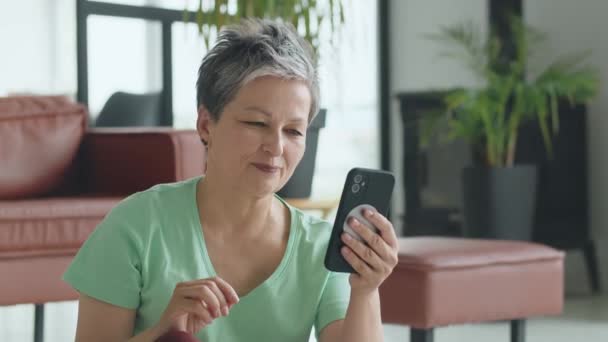 老妇人在家里练完瑜伽后打电话给她的朋友 成熟的女人有积极的体育生活 她在智能手机上有视频通话 高质量的4K镜头 — 图库视频影像