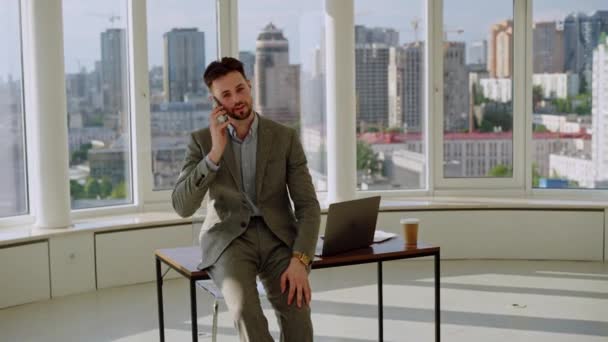 年轻的经理老板是一位专业的生意人 他坐在办公室的桌子上 看着智能手机屏幕 打电话 有大办公室和城市背景 高质量4K — 图库视频影像