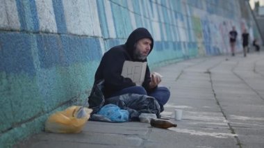  Hayatta kalmak için para isterken yardım masasıyla şehirde yerde oturan evsiz genç adam. Eski, sağlıksız yemekleri yemek. Yüksek kalite 4k görüntü