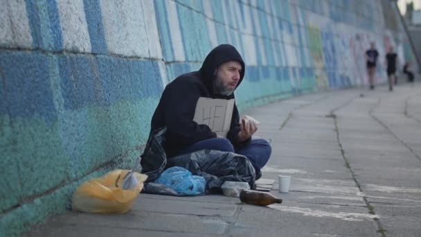  бездомный бедный молодой человек, сидящий на земле в городе со столом Помощи, прося денег, чтобы выжить. Есть старую вредную пищу. Высококачественные 4k кадры