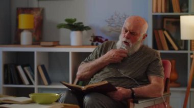 Nefes maskeli yaşlı adam sandalyede otur ve kitap oku. Yaşlı adam zor nefes aldığı için nebulizör kullanıyor. Çeşitli akciğer hastalıklarının tedavi teması. Yüksek kalite 4k görüntü