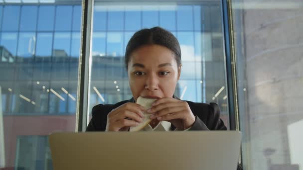 若いアフロ系アメリカ人の女性は 片手で何かをタイプし 別のものと一緒に食べます オフィスの黒人女性が朝食にサンドイッチを食べるのは時間がないからだ ストレスの多い仕事と不健康な食事 — ストック動画
