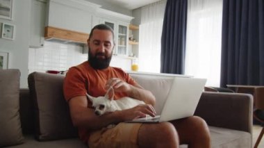 Yetişkin bir adam tek eliyle dizüstü bilgisayarda çalışırken aynı zamanda sevimli köpeğini okşar. Evcil hayvanıyla internetten çalışan serbest çalışan bir adam. Yüksek kalite 4k görüntü