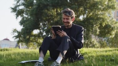 Genç adam parkta tablet karıştırıyor. Pozitif işadamı bütçe cüzdanını kontrol ediyor. Adam boş zamanını dışarıda harcıyor ve mükemmel bir sevgili bulmak için flört uygulamasına takılıyor. Yüksek kalite 4k görüntü