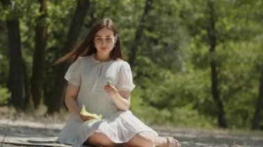Olumlu duygularla gülümseyen mutlu bir kız yaz ormanında kumların üzerinde oturur. Saçlı, rüzgarda esen beyaz elbiseli genç bir bayan neşeyle inceler ve parlak bir görüntü sergiler.