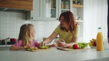 Mutlu bir anne ve kızı mutfak masasında yüzlerinde gülümsemeyle yemek tariflerini tartışıyor ve sandviç hazırlıyorlar. Basit ve sağlıklı yiyeceklerin gösterimi. Şey...