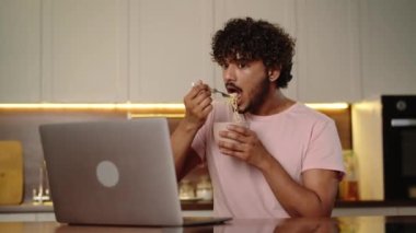 Fazla mesai yapan genç adam evde ramen çorbası içiyor. Komik adam elinde hazır erişteyle bilgisayarda video izliyor. Erkek, yemeğinin tadını çıkarıyor. İzleme serisi. Geç saatte akşam yemeği. Yüksek