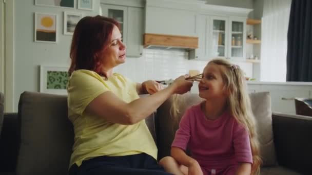 フレンドリーな笑顔の母親は 娘と一緒に部屋のソファーに座り マットニーの準備のために髪を編みます 若い女性が子供にヘアスタイルのファッションのトレンドについて話します イギリス — ストック動画