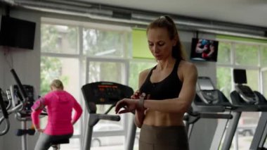 Spor salonundaki genç bir kız sporcu antrenman için hazırlanıyor. Bir kadın, fiziksel aktivitenin nabzını ve yönetmelik yoğunluğunu kontrol etmek için fitness saatini dikkatlice özelleştirir. Sağlıklı olma kavramı