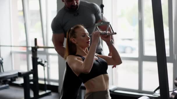 在男子健身教练的指导下 一个女孩在体育馆里做运动 教练支持她的后背 女人用起吊拉索做运动 灵活地举起和伸展武器 — 图库视频影像