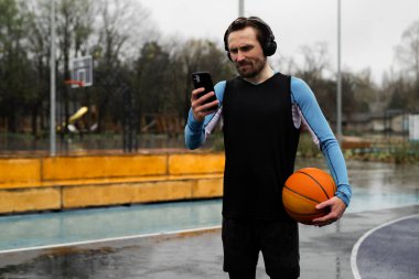 Müzik kulaklıklarını değiştirmek için akıllı telefon kullanan amatör basketbolcu kavramı. Güçlü sporcu, kablosuz kulaklıkları dinle ve topu tut. Profesyonel atlet spor alanında telefonu tutuyor. Yapıyor