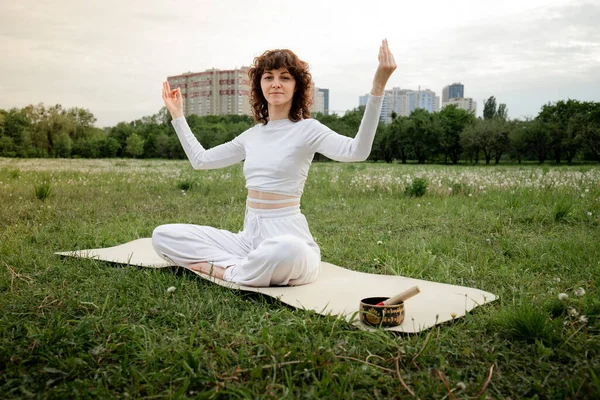Concept of calm sportswoman meditation. Breathing techniques on nature background. Young lady yoga therapy practice. Woman sitting caremat raised hands up in lotus posture. High quality photo