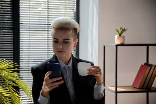 年轻的女职员在使用智能手机的时候有喝咖啡休息时间 由于有了新的现代电话 女企业家可以与人聊天了 女商人用一杯浓缩咖啡和移动饮料打发时间 — 图库照片