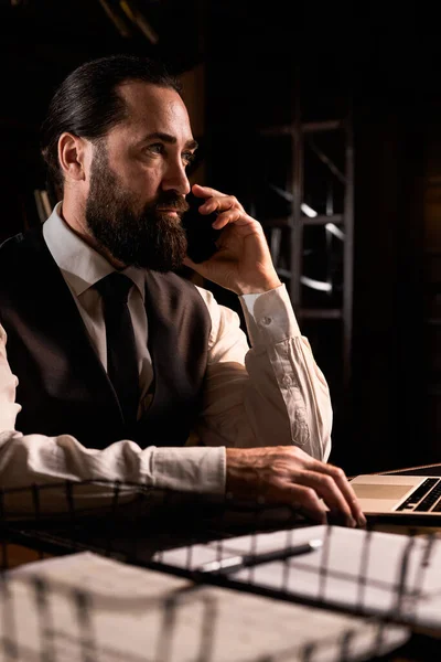 Successful investor talking on smartphone. Businessman works in late night at his home office. Man making phone call when browsing laptop. Entrepreneur has urgent online discussion overtime. Elegant