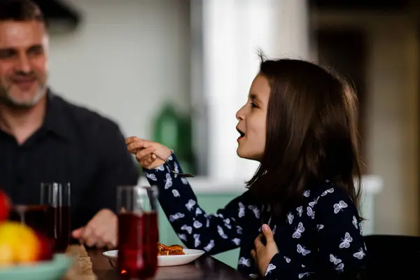 Schoolgirl eating pasta bolognese on the kitchen table. Cute daughter blowing hot fork with food. Small girl enjoying her meal with father. Christians family lunch. Family talking at breakfast. Kid