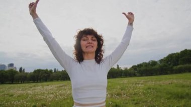 Beyaz eşofmanlı genç bir kadın yoga eğitiminde ellerini kaldırıyor. Sporcu kadın, Pranayama meditasyonu üzerine yoğunlaşmıştı. Kız parkta elini kaldırır ve zihnini açar. Yüksek kalite 4k görüntü